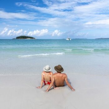 A couple sitting on the sand in the water looking out an an island and a catamaran