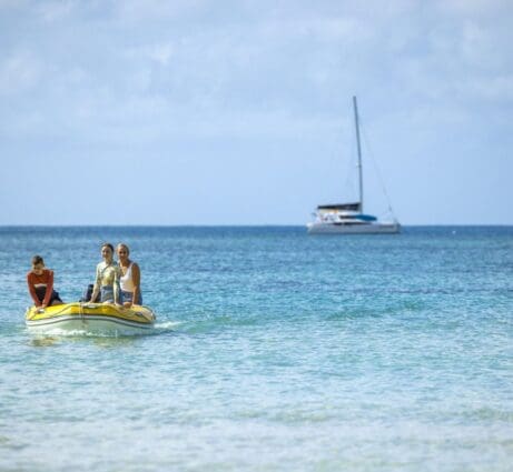 3 people in a yellow dinghy, with a sailing catamaran in the background.