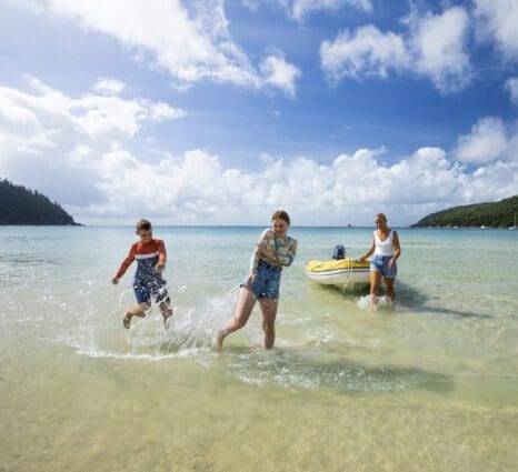 Two children playing in the water on a beach whilst a woman pulls a boat onto the beach.